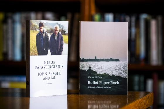 Winners of this year's National Biography Award. The photo shows the cover of two books standing upright. The cover on the left depicts two people walking side by side outdoors, and the one on the right depicts a view looking out to the water with the city in the background.