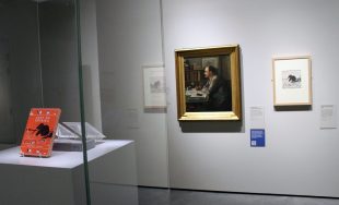 A painting of a man in profile. There is an orange book, 'Just So Stories' in a glass case and some other documents next to the painting.