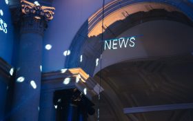 A neon sign with the text ‘NEWS’ hanging on a thin pole in front of an arched doorway. There are soft blue and purple lights illuminating the space.