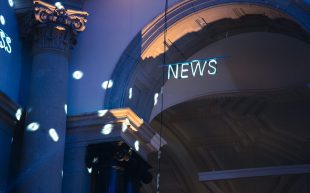 A neon sign with the text ‘NEWS’ hanging on a thin pole in front of an arched doorway. There are soft blue and purple lights illuminating the space.