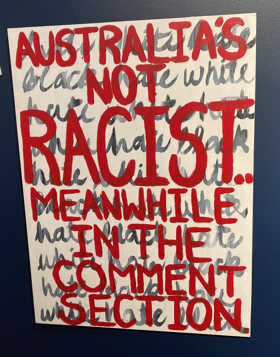 A white board with grey cursive writing on it, overwritten with big red letters saying AUSTRALIA'S NOT RACISE MEANWHILE IN THE COMMENT SECTION. Teneille Nuggins, 'Behind the Surface' (2025), CIAF 2025 Art Fair Showcase, installation view.