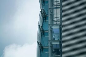Two people stand behind the glass wall of an external elevator in this photograph, taken at a distance, which also shows the horizontal lines of the building's cladding, and the sharp lines of the building's edge against a cloudy sky. The photo illustrates ArtsHub's On the mov column, a weekly round-up of arts sector appointments across Australia.