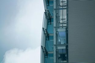 Two people stand behind the glass wall of an external elevator in this photograph, taken at a distance, which also shows the horizontal lines of the building's cladding, and the sharp lines of the building's edge against a cloudy sky. The photo illustrates ArtsHub's On the mov column, a weekly round-up of arts sector appointments across Australia.