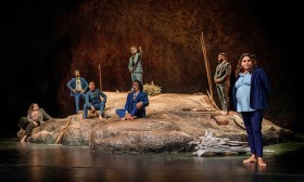 The cast of The Visitors - Seven people in suits either sitting or standing on a rock-like formation.