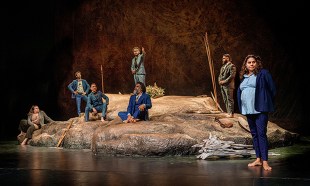 The cast of The Visitors - Seven people in suits either sitting or standing on a rock-like formation.