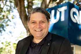 Wesley Enoch, a highly respected cultural leader and First Nations man, smiles at the camera. He is wearing black and a QUT sign is visible behind him.