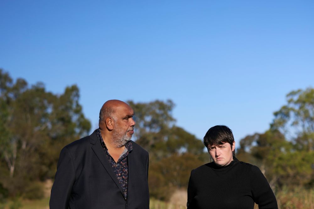 Kutcha Edwards (left) and Cash Savage (right) will perform together at this year's Junction Arts Festival in Launceston. The photo shows an older, dark-skinned Aboriginal man with a shaven head, and an androgynous, fair-skinned woman with dark hair squinting as she looks into the sun and towards the camera. The Australian bush is visible behind them.