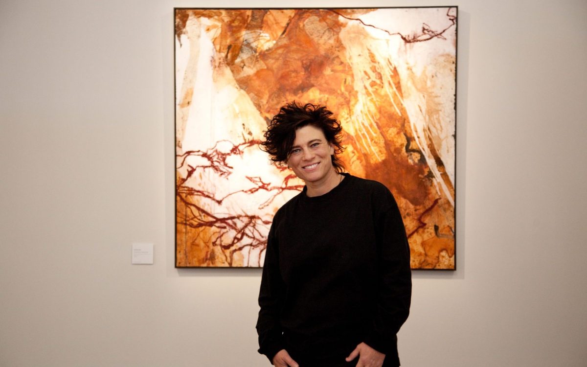 Sophie Cape, an ex-athlete, wearing all black with a big smile, standing in front of abstract landscape painting in rusty colours.