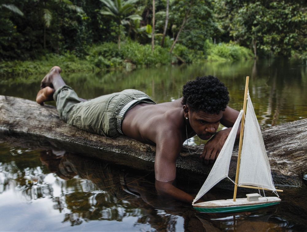 Naomi Hobson, 'Present & Beyond' 2024, at the 2025 Telstra NATSIAA. Hobson's award-winning photograph shows a shirtless 16 year old Aboriginal youth, Dallas, lying on a log and playing with a toy yacht on the Coen River.