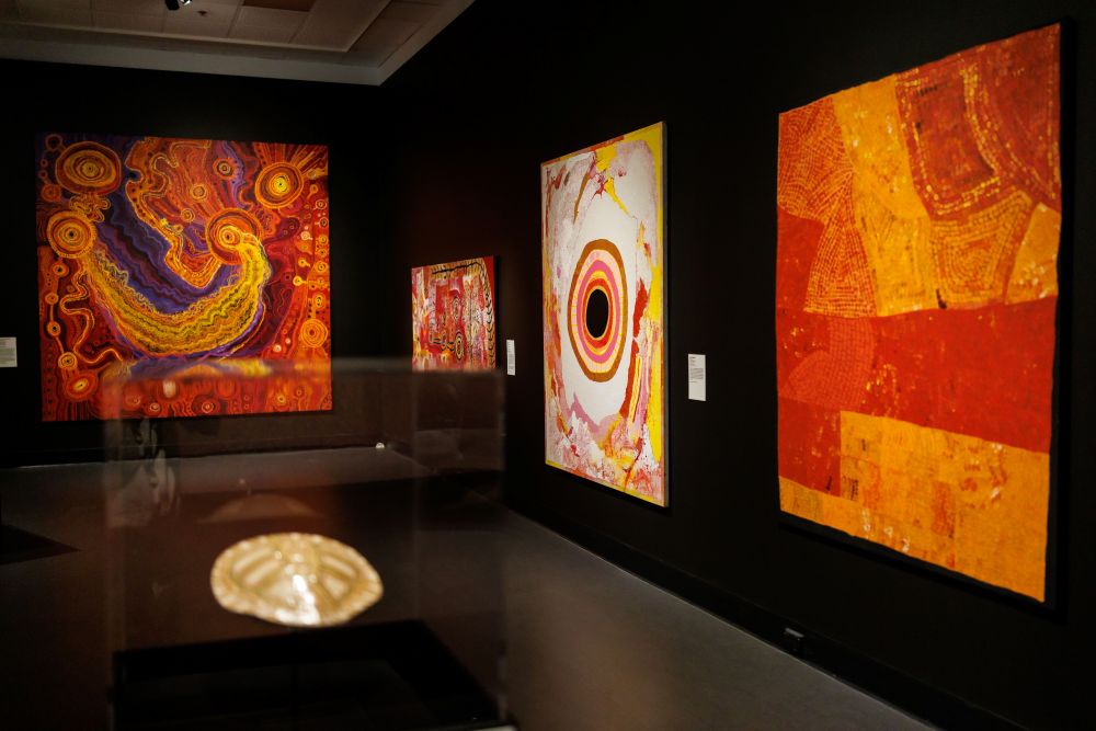Installation view, the 2025 Telstra NATSIAA at MAGNT, Darwin. A range of striking and colourful artworks hanging on the black walls of the Gallery.