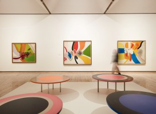 Gallery view of an exhibitio with colour abstract paintings and round table sculptures. Janet Dawson
