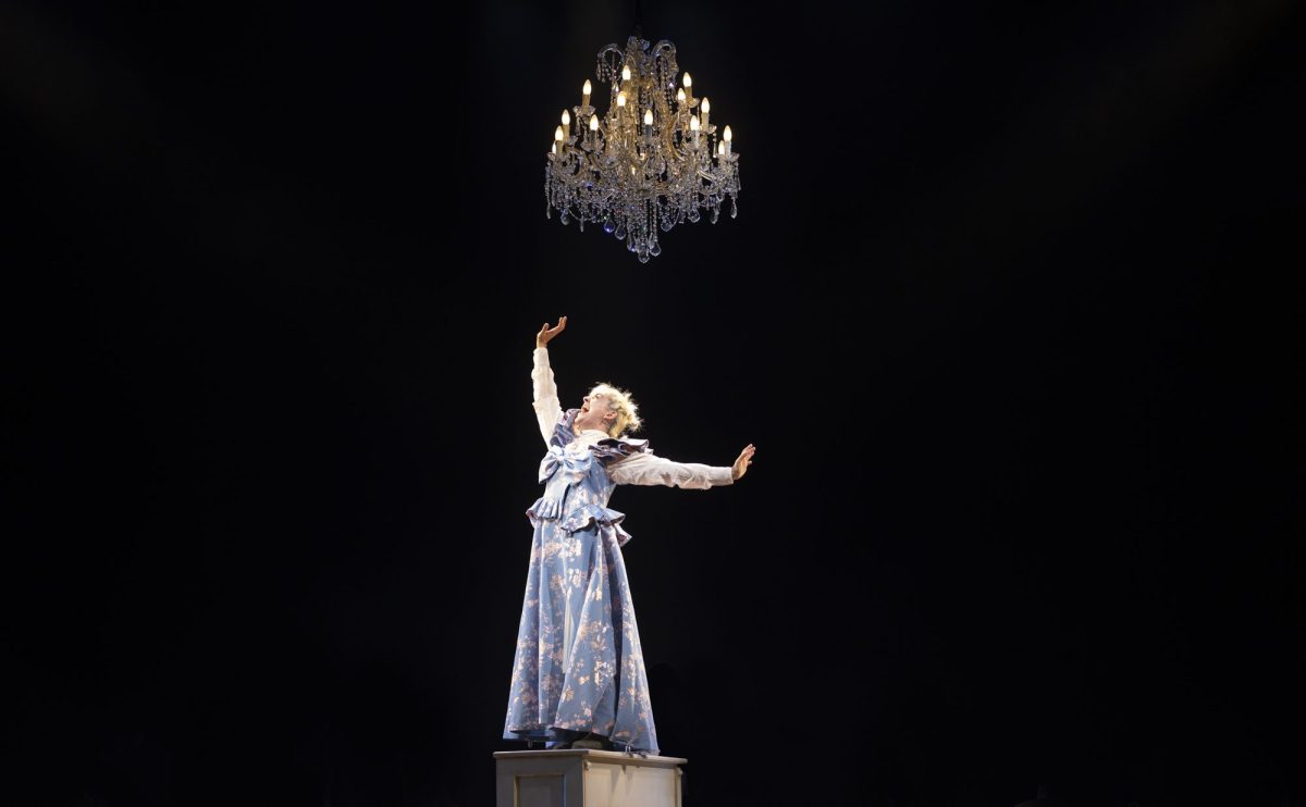 Kala Gare in MTC's original stage musical 'My Brilliant Career' which returns for an encore in the company's 2026 season. The photo shows a fair haired and fair skinned young woman in period dress posing dramatically beneath a glittering chandelier. Her head is tiled back and her eyes closed as she sings, her right arm thrusts upward and her left arm swings back behind her.