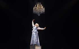 Kala Gare in MTC's original stage musical 'My Brilliant Career' which returns for an encore in the company's 2026 season. The photo shows a fair haired and fair skinned young woman in period dress posing dramatically beneath a glittering chandelier. Her head is tiled back and her eyes closed as she sings, her right arm thrusts upward and her left arm swings back behind her.