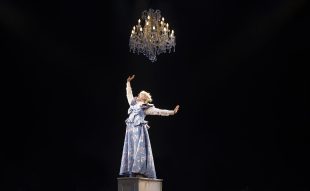 Kala Gare in MTC's original stage musical 'My Brilliant Career' which returns for an encore in the company's 2026 season. The photo shows a fair haired and fair skinned young woman in period dress posing dramatically beneath a glittering chandelier. Her head is tiled back and her eyes closed as she sings, her right arm thrusts upward and her left arm swings back behind her.