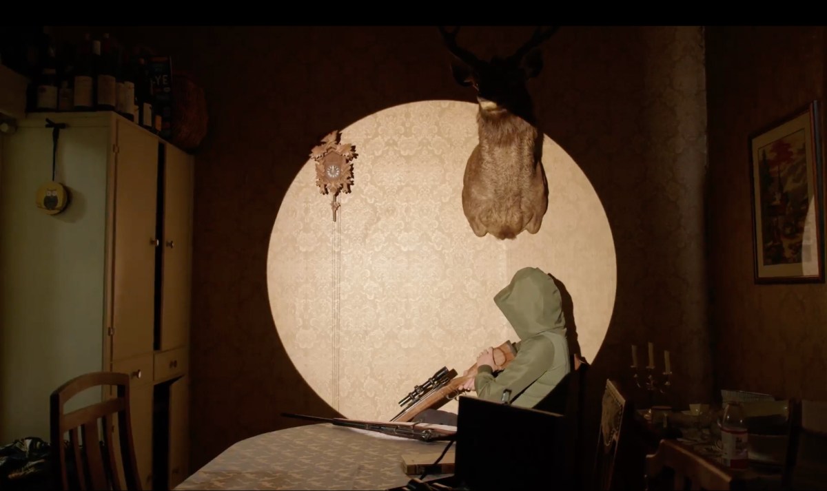 A figure in a domestic dining room holding a rifle with a round spotlighting shining on them. Free exhibitions in Tasmania.