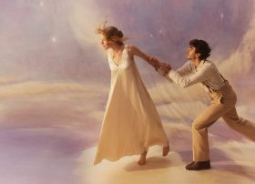 Promotional image for Opera Australia's 2026 production of 'La Bohème'. The photo shows a young woman in white walking away from a young man, who clings to her left hand as if imploting her to stay.