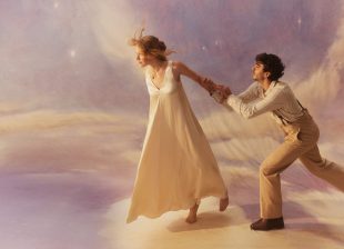 Promotional image for Opera Australia's 2026 production of 'La Bohème'. The photo shows a young woman in white walking away from a young man, who clings to her left hand as if imploting her to stay.
