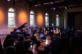 ANAM students performing at Abbotsford Convent. The atmospheric photograph, taken in a historic venue with picturesque windows and a stage visible in the background, shows a seated audience listening to a group of six musicians, including strings, woodwind and a piano; one of the students stands behind a computer screen, suggesting the presence of electronics in the score being performed.
