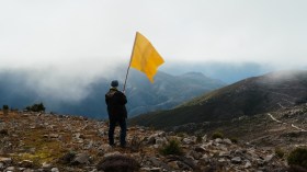 Man standing in foggy mountains holding up a yellow flag. The Unconformity