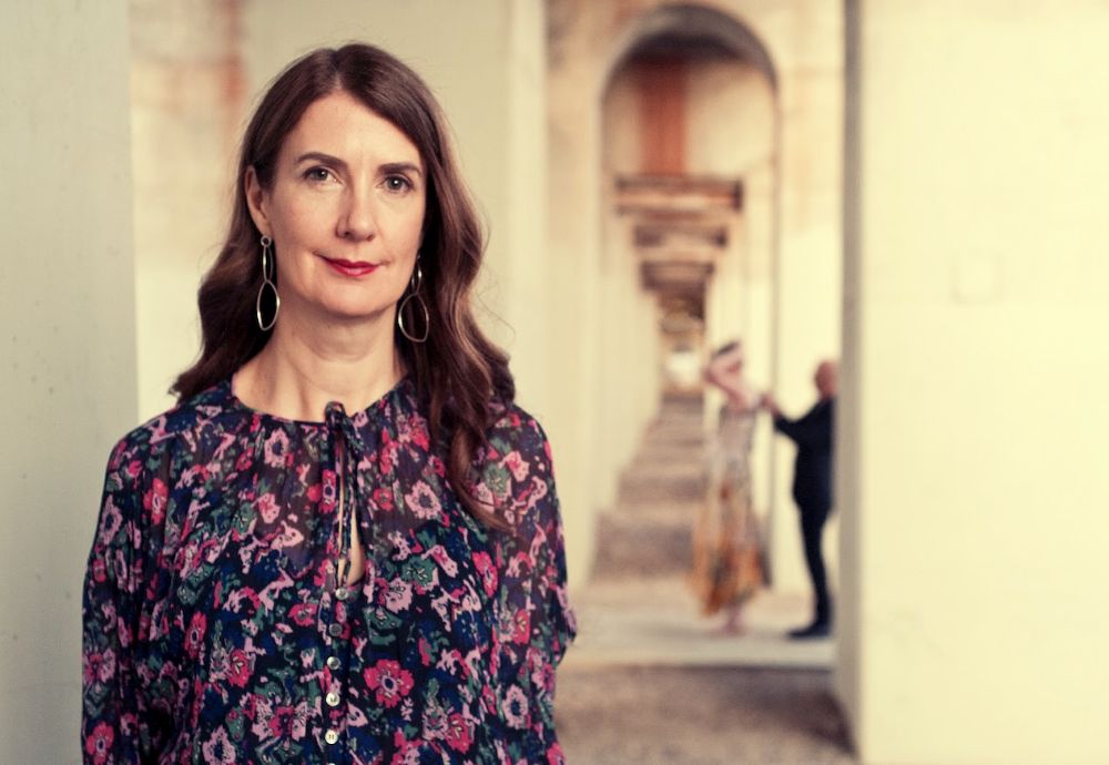 ANAM's next Artistic Director Anna Goldsworthy, a woman with long brown hair and silver earings, and wearing a floral dress tied at the neck. She stands in a portico, the arches of which are blurred in the distance behind her. The photo is included as part of ArtsHub's coverage of national arts sector appointments, On the move.