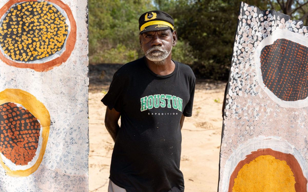An Aboriginal man standing between two large bark paintings on the beach. Free exhibitions in the Northern Territory.