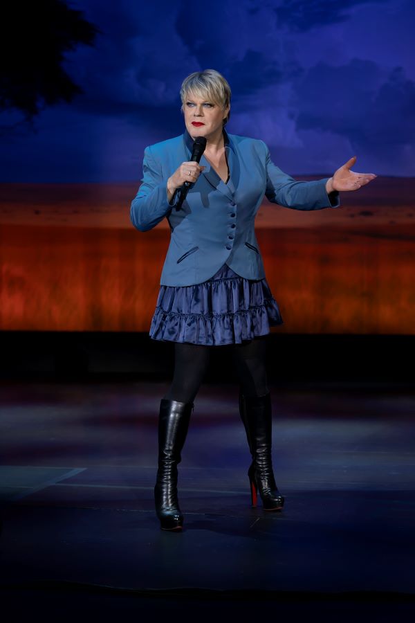 Eddie Izzard brings ‘Eddie Izzard – The Remix Tour Live’ to Australia in November-December this year. The photo depicts Eddie Izzard performing on stage: she is a fair-skinned, middle aged woman holding a microphone in her right hand and gesturing with her left. Eddie wears a blue jacket and a ruffled blue skirt in a darker shade, black tights and long black boots. She has short blonde hair and is wearing makeup and lipstick.