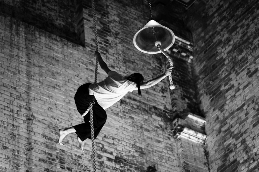 Circus artist Bridie Hooper will perform 'For Who the Bell Tolls' at Junction Arts Festival 2025. The black and white photograh shows a barefoot circus artist dressed in black pants and a white t-shirt and presumably mid-routine, suspended from a rope looped around their legs. The artist hangs in a bell tower, grasping a second rope attached to the clapper of a large bell, which they look ready to ring.