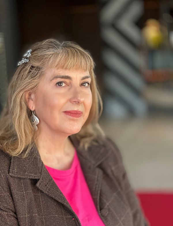 Gail Harradine is the inaugural First Nations Representative on the Public Galleries Association of Victoria Board. The photograph depicts a fair-skinned Aboriginal woman looking calmly at the camera. She wears a brown jacket over a pink shirt, and a silver earring is visible under her dyed blonde hair. The photograph illustrates ArtsHub's On the move column of Australian arts sector appointments.