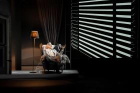 A man, Ian Stenlake, sitting in a chair near a shuttered blind in The 39 Steps.