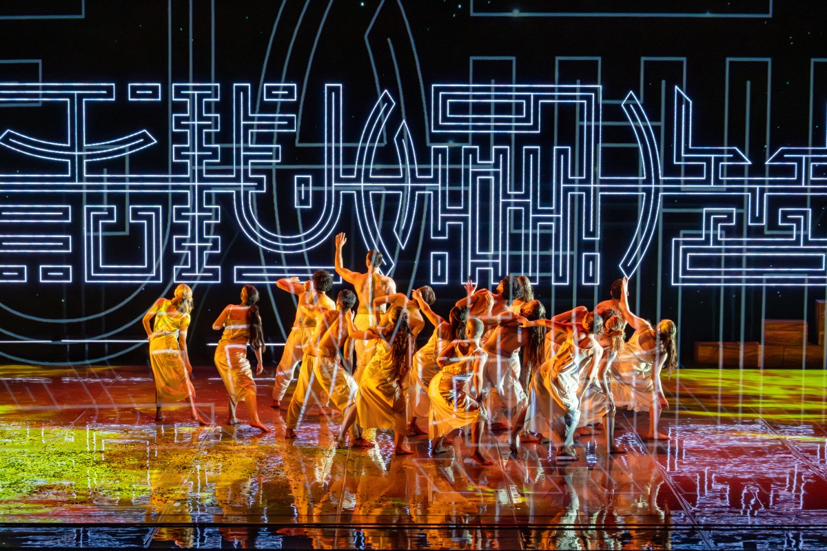 A group of 16 dancers tightly clustered on stage facing the same direction while yellow and orange light illuminate their bodies. A projection of symbols are overlaid onto the stage. Bangarra rehearsal director shares insights. 