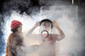 A publicity image for 'Influencer'. a Junction Arts Festival dance production about social media. The image shows a group of people half-visible in a cloud of theatrical smoke: a pasted-on photo of a blonde-haired woman obscures one of the faces, which has a male figure in a brown coat and red beanie holding up a ring-light in front of them.