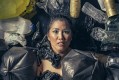 A promotional image for La Boite and Dead Puppet Society's climate change production, 'We're All Gonna Die!'. The photo shows a female Asian-Australian actor's shocked looking face as they are engulfed by spreading rubbish and garbage.