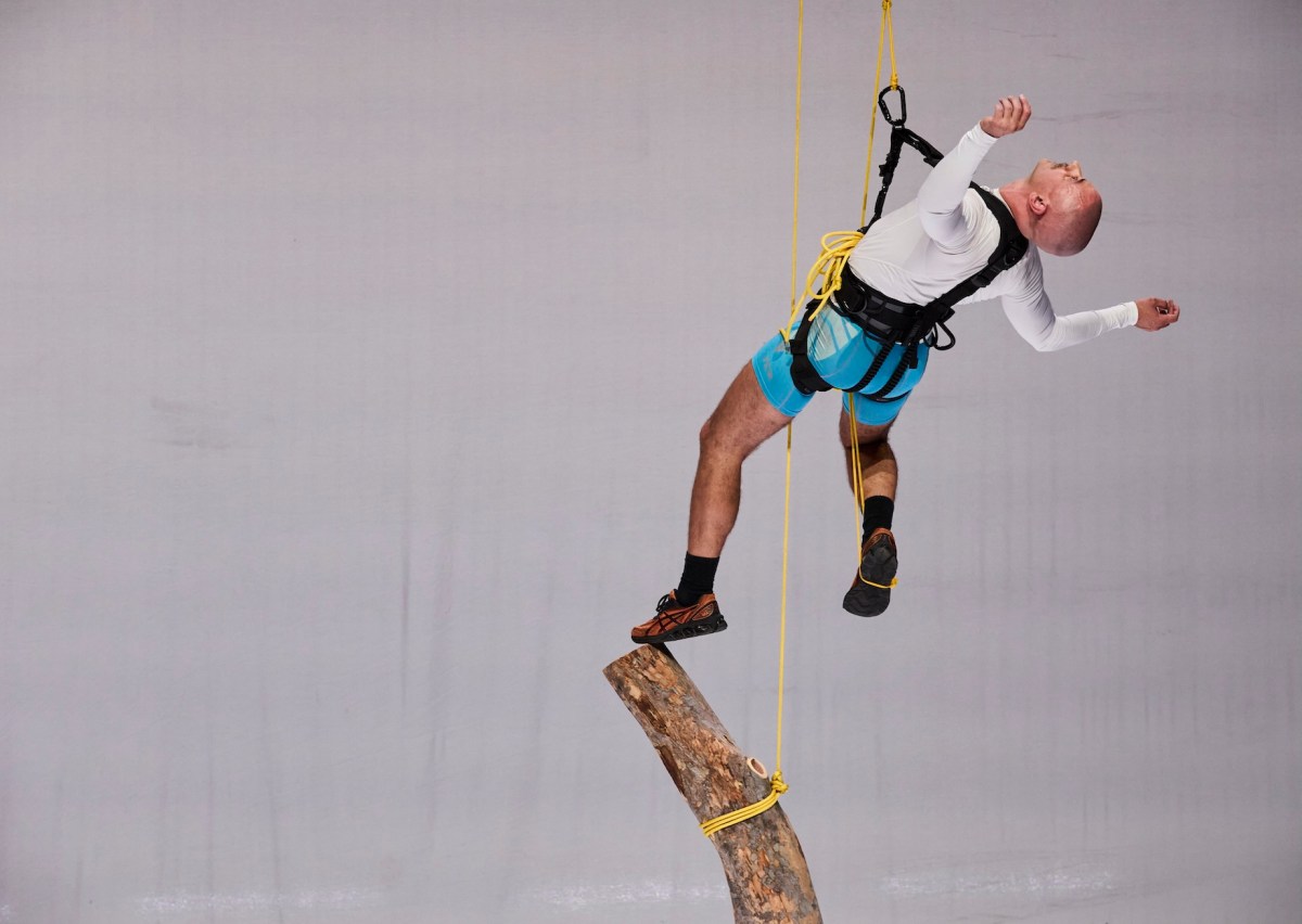 Man suspended by climbing harness with a log hanging beneath him. The Unconformity