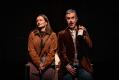 A man and a woman sitting next to one another. He's holding up a wine bottle. Sarah Morrison and Bobby Fox in Ordinary Days.