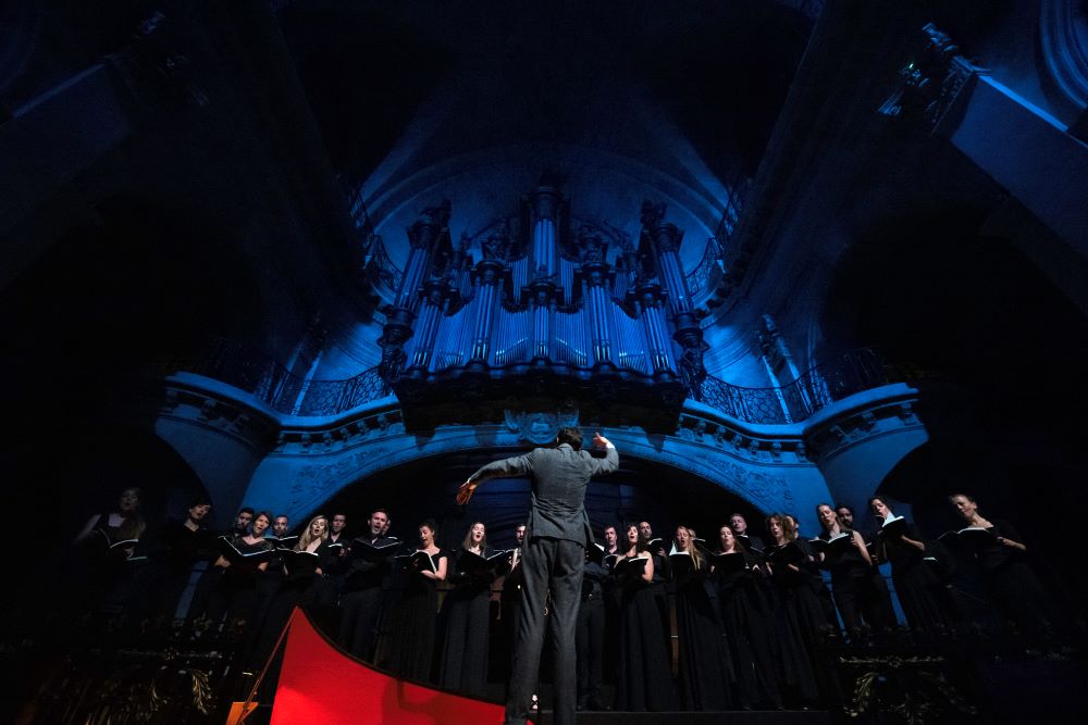 France's Ensemble Pygmalion will perform exclusively at the 2026 Adelaide Festival. Photo: Fred Mortagne. The photograph, taken from a low angle, depicts a choirmaster vigorously conducting the Ensemble's choir, who stand arrayed before him.