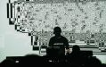 Ryoji Ikeda with wearing black with his face hidden under a cap, standing in front of a glitchy digital background with his laptop.