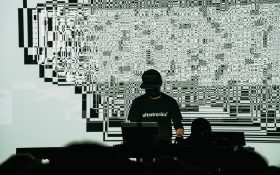 Ryoji Ikeda with wearing black with his face hidden under a cap, standing in front of a glitchy digital background with his laptop.