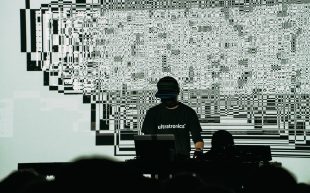 Ryoji Ikeda with wearing black with his face hidden under a cap, standing in front of a glitchy digital background with his laptop.
