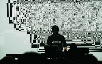 Ryoji Ikeda with wearing black with his face hidden under a cap, standing in front of a glitchy digital background with his laptop.