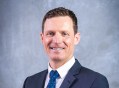 A profile photo of a man with brown hair, a dark suit and blue tie. He is smiling. Michael Sterzinger, CEO of Queensland Symphony Orchestra. QSO