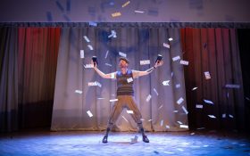 Paper notes flying onto a stage with one performer standing in the centre with arms outstretched.