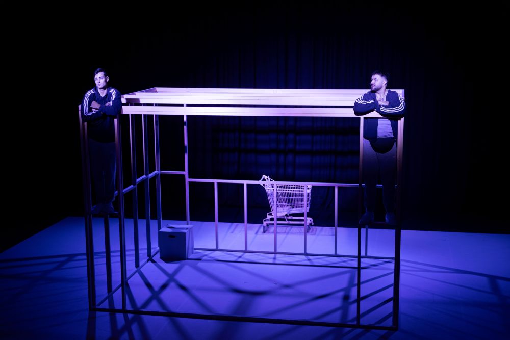 A scene from Mudlark Theatre's 2024 production 'The Box', featuring (L-R) actors Nelson Clay and Corey Saylor-Brunskill. Mudlark is one of the initial members of the Tasmanian Live Performance Exchange. The blue-lit photo shows two male-presenting performers in dialogue; they have climbed the bare frame of a unit or small house under construction.