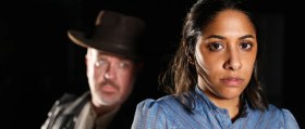 A publicity image for The Street Theatre's production of 'The Chosen Vessel', a new Aboriginal Gothic play by Dylan Van Den Berg. The photo depicts an Aboriginal woman staring at the camera; a white man stands ominously behind her, disappearing into the shadows.