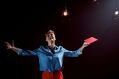 A scene from 'The Past is a Wild Party' by Noelle Janaczewska, one of the 7-ON playwrights. The photo depicts a female-presenting actor with blonde hair and wearing a long-sleeved blue shirt and red trousers. Her arms are outstretched, her head tilted back and her mouth open as if she is exclaiming ecstatically. She holds a closed book with a red cover, perhaps a play script, in her left hand.