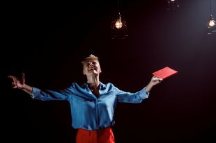 A scene from 'The Past is a Wild Party' by Noelle Janaczewska, one of the 7-ON playwrights. The photo depicts a female-presenting actor with blonde hair and wearing a long-sleeved blue shirt and red trousers. Her arms are outstretched, her head tilted back and her mouth open as if she is exclaiming ecstatically. She holds a closed book with a red cover, perhaps a play script, in her left hand.