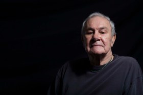 A promotional photograph of the late Noel Tovey AM from the 2014 season of 'Little Black Bastard', presented by ILBIJERRI and Arts House. The photo shows an elder but still vigorous Aboriginal man with grey hair; he poses regally and is looking gravely down at the camera.