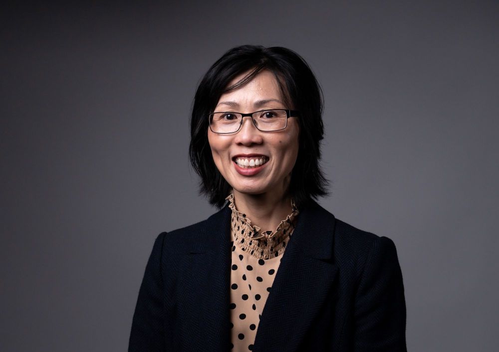 Vivienne Nguyen AM has joined the Abbotsford Convent Foundation Board. The photo depicts an older Vietnamese-Australian woman smiling at the camera against a grey backdrop. She has short, black hair and wears thin, black-framed glasses, a black-spotted brown blouse with a ruffled neck, and a black jacket.