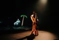 A scene from the 2025 ROOKE production 'Wilds', featuring performers Conor Wild and Freyja Wild. Contemporary circus company ROOKE is one of the inaugural members of the Tasmanian Live Performance Exchange. The photograph depicts a female-presenting acrobat wearing brown garments and performing a headstand; in the background, a male-presenting performer in blue clothes and black boots sits in a folding chair beside an inflatable palm tree.