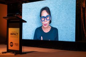 Rhoda Roberts accepting the Wendy Blacklock Award at PAC Australia's Impact Awards. The photo shows an older Aboriginal woman with blue-framed glasses, black hair and wearing red lipstick and a dark blue top, speaking on video against a light blue textured background.