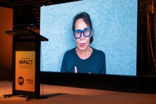 Rhoda Roberts accepting the Wendy Blacklock Award at PAC Australia's Impact Awards. The photo shows an older Aboriginal woman with blue-framed glasses, black hair and wearing red lipstick and a dark blue top, speaking on video against a light blue textured background.
